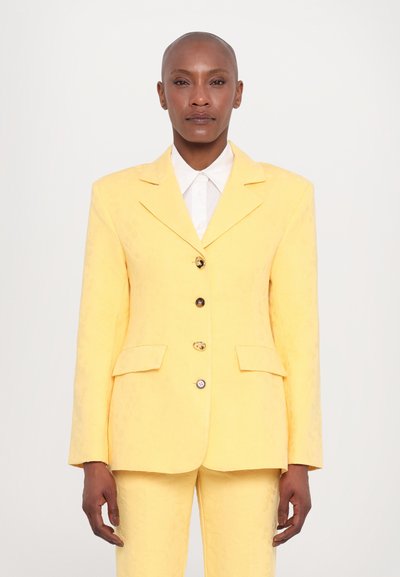 Blazer jaune sur mesure en tissu texturé avec un revers cranté, à boutonnage simple, quatre boutons et deux poches avant. Chemise blanche à col en dessous.