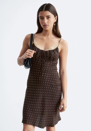 Mujer con vestido marrón de lunares con tirantes finos y ribete de encaje, que lleva un pequeño bolso negro de hombro y lleva joyas doradas.