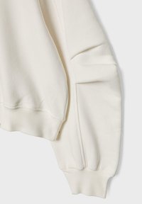 Des manches de sweat-shirt de couleur crème avec des poignets côtelés et des détails de couture subtils sur un fond gris clair.