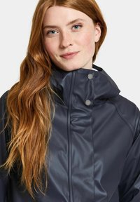 Marinblå vattentät jacka med hög krage, tryckknappar och mjuk, glänsande yta. Närbild på ansikte med långt, vågigt hår.