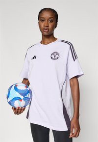 Bílý nadměrný tričko s logem Manchester United, černými pruhy na rukávech, doplněné barevným vzorovaným fotbalovým míčem drženým v ruce.