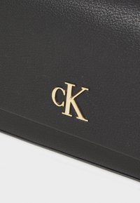 Juodas odinis krepšys su tekstūriniu paviršiumi, išryškintu auksiniu "cK" logotipo akcentu. Paprastas, stačiakampis dizainas su švariomis linijomis.