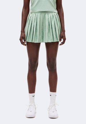 Kobieta ubrana w jasnozieloną plisowaną spódnicę, w połączeniu z białymi skarpetkami Nike i białymi butami sportowymi, stoi na jednolitym tle.