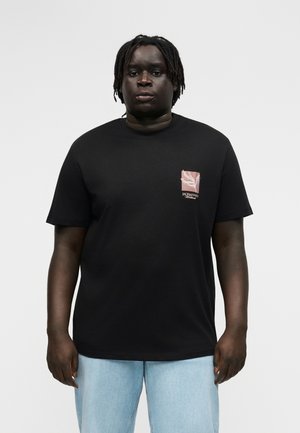 Rövid dreadlock hajjal rendelkező férfi, fekete pólót visel, melynek mellkasán kis rózsaszín virágos logó található, valamint világoskék farmert, fehér háttér előtt áll.