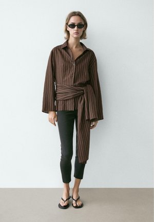 Femme portant des lunettes de soleil foncées, une chemise cache-cœur à manches longues rayée marron, un pantalon noir court et des sandales noires, debout contre un mur uni.