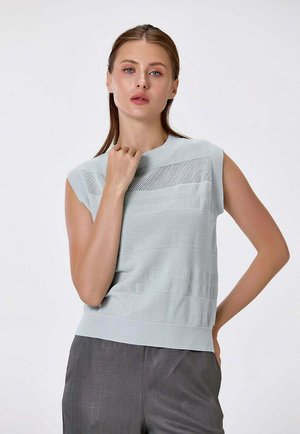 Jeune femme aux cheveux châtain clair portant un top en maille texturée bleu clair sans manches et un pantalon gris, posant avec la main près de la clavicule.