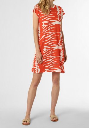 Kobieta w pomarańczowo-białej sukience o krótkich rękawach, długości do kolan, w beżowych sandałach na wsuwkę, stojąca na jednolitym tle.