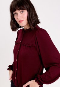 Bordeauxfarbene Bluse mit Knopfleiste aus strukturiertem Stoff, mit Rüschendetails am Kragen und an der Brust sowie puffärmeln mit elastischen Bündchen.