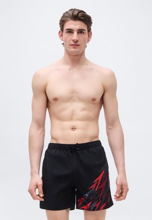 Nuori mies lyhyillä hiuksilla, joka pukeutunut mustiin uimashortseihin, joissa on punaisia ja harmaita salamasomisteita, seisomassa yksinkertaisen vaalean taustan edessä.