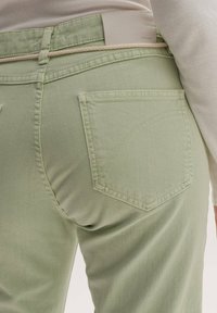 Jeans in denim verde chiaro con una tasca posteriore caratterizzata da cuciture curve. Il girovita ha un cinturino chiaro a contrasto.