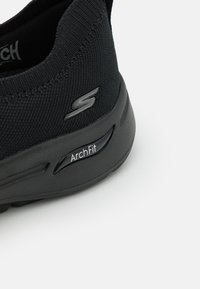 Zapatilla deportiva negra con upper de punto elástico, que presenta el logo "S" y el branding "ArchFit" en el lateral. Suela ligera, diseño elegante.