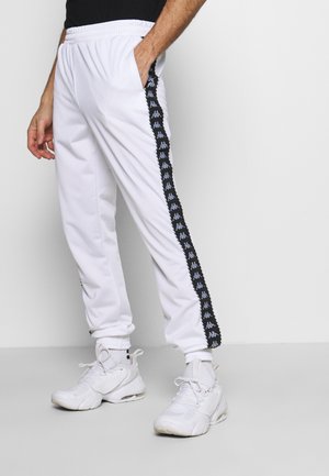 Witte sportbroek met zwarte zijstrepen met daarop een herhaald logo-ontwerp. Elastische tailleband en omgeslagen pijpen. Draagt witte sneakers.