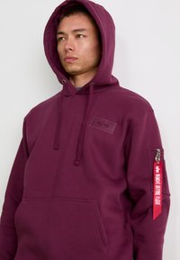 Haut à capuche bordeaux avec une poche avant, des cordons de serrage et une poche à fermeture éclair sur la manche comportant une étiquette rouge avec le texte "ENLEVEZ AVANT LE VOL".