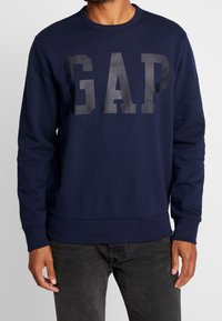 Muž nosící tmavě modrou mikinu s velkými tmavými písmeny "GAP" na hrudi a černé džíny, stojící na jednobarevném pozadí.