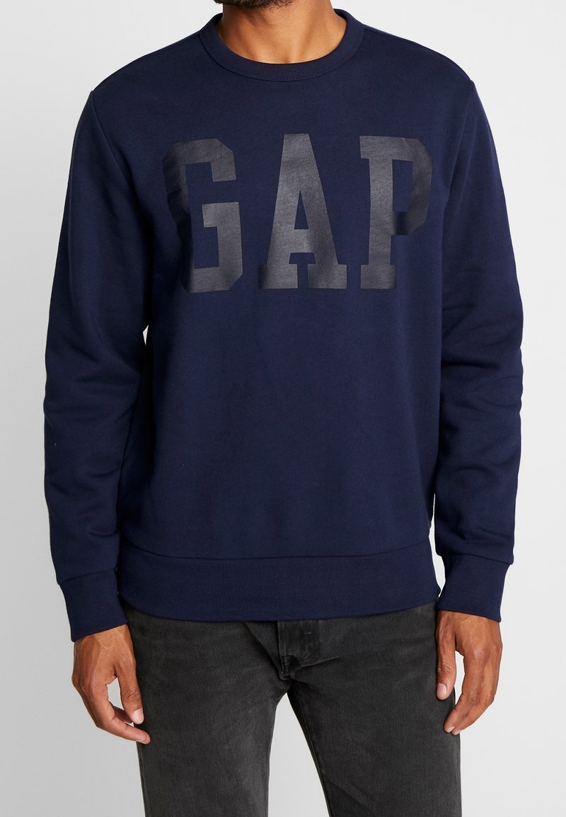 Muž nosící tmavě modrou mikinu s velkými tmavými písmeny "GAP" na hrudi a černé džíny, stojící na jednobarevném pozadí.