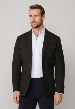 Dunkel kariertes Sakko mit slim Fit, zwei Knöpfen, Brusttasche und braunen Knöpfen, kombiniert mit einem weißen Hemd und navy Hosen.