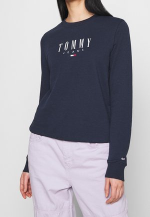 Kvinde iført en marineblå Tommy Jeans sweatshirt med hvidt logotekst og lyse lavendelfarvede bukser, stående mod en ensfarvet baggrund.