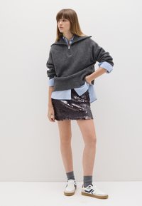 Suéter gris oscuro con cremallera y cuello alto, superpuesto a una camisa de rayas azul claro y una mini falda brillante de lentejuelas. Zapatillas blancas con detalles en gris completan el look.