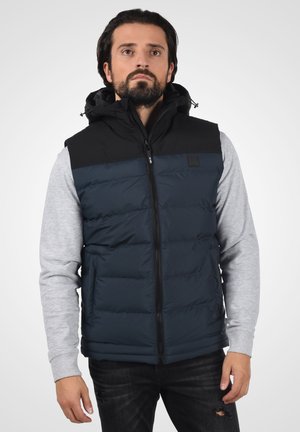 Gilet imbottito blu navy con cappuccio e spalle nere, dotato di zip frontale e tasca con zip, indossato su una maglietta a maniche lunghe grigia.