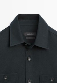 Camicia nera in tessuto strutturato con colletto a punta, taschino e bottoni neri. Presenta un'etichetta con la scritta "Massimo Dutti" e prodotta in Cina.