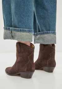 Stivaletti alla caviglia in suede marrone con un design a cucitura curva e tacco a strati, abbinati a jeans in denim arrotolati con un risvolto blu chiaro.