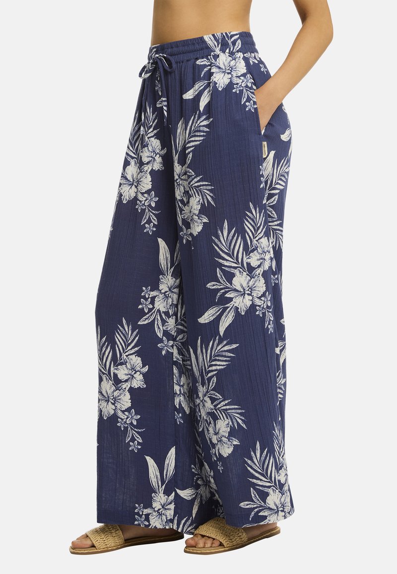 Model in een vloeiende, marineblauwe broek met een witte bloemenprint en zakken, gecombineerd met lichtbruine sandalen. Stijlvolle zomerse outfit.