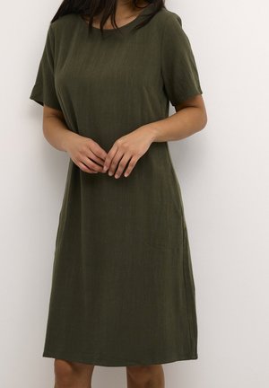 Woman wearing a simple knee-length olive green short-sleeve dress standing against a white wall with hands loosely clasped.