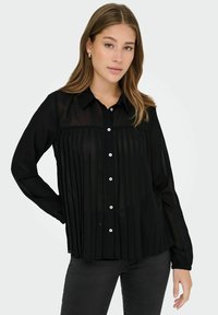 Černá button-up košile s vertikálními sklady, průhlednou tkaninou a klasickým límcem. Obsahuje kontrastní knoflíky a volný střih.