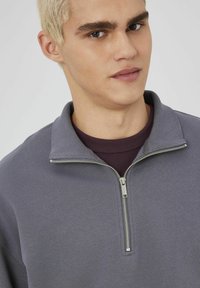 Sweatshirt gris zippé avec un col replié et un tissu texturé, dotée d'une fermeture éclair argentée et d'un t-shirt bordeaux superposé en dessous.