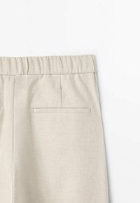 Beige pellava-housut joustavalla vyötäröllä, joissa on yksi takatasku ja sileä rakenne. Muotoilu on minimalismissia ja toiminnallista.