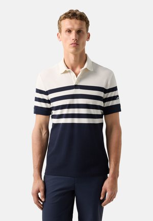 Man draagt een poloshirt met korte mouwen en witte en marineblauwe horizontale strepen, en een marineblauwe broek, staand tegen een effen achtergrond.
