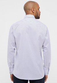 Camicia bianca a maniche lunghe con motivo a rombi blu, colletto polo e orlo arrotondato. La vista posteriore mostra maniche lunghe e una finitura curata.