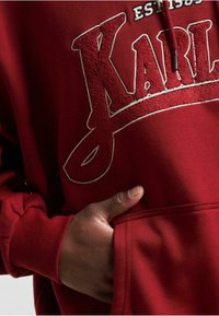 R�ød hettegenser med teksturert "KARL"-logo i hvit brodering, en frontlomme i kenguru-stil og ribbede mansjetter og nederkant, med myk stofffølelse.