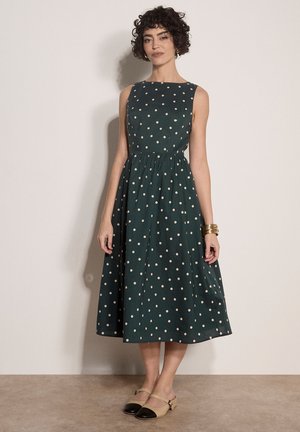 Vrouw met kort krullend haar die een mouwloze donkergroene midi-jurk met witte stippen en beige platte schoenen met zwarte neus draagt.