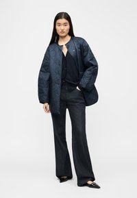 Donna che indossa una giacca trapuntata nera sopra una maglia nera, pantaloni larghi scuri e scarpe nere con punta a punta, in piedi con una mano in tasca.