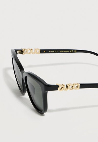 Μαύρα ορθογώνια γυαλιά ηλίου με σκοτεινούς φακούς, διακοσμημένα με χρυσές λεπτομέρειες λογοτύπου Gucci στα πλαϊνά και κείμενο "Κατασκευάστηκαν στην Ιαπωνία" στο εσωτερικό του βραχίονα.