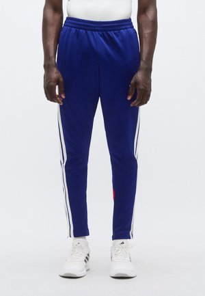 Pantalones deportivos azules con franjas blancas laterales, cintura elástica y un detalle rojo. La tela parece suave y ligera. Combinados con zapatos blancos.