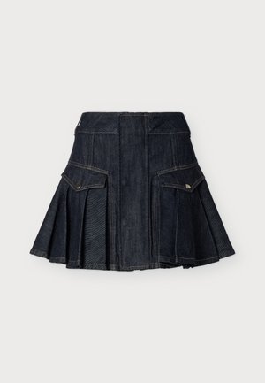 Mujer que lleva una blusa blanca de manga larga y hombros al descubierto, falda mini plisada de mezclilla oscura con bolsillos y botas altas de cuero marrón, posando frente a un fondo liso.
