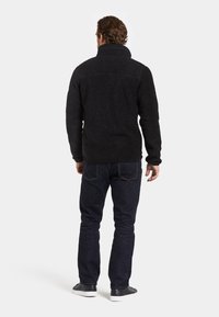 Svart fleecepully med hög krage, texturerad yta och lång ärm, kombinerad med mörk denimjeans och svarta skor.