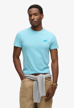 T-shirt in cotone azzurro chiaro a maniche corte con collo tondo e un piccolo logo sulla parte sinistra del petto; abbinato a pantaloni beige e un maglione grigio annodato in vita.