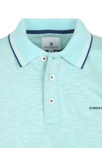 Lichtblauw poloshirt met een textuurstof, een knoopsluiting met drie knopen, en contrasterende marineblauwe kraag en manchetten met gestreepte details.