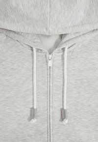 Sivi zip-up dukserica napravljena od mekanog, strukturiranog materijala s kapuljačom, koja ima bijele vrpce za zatezanje i prednji zatvarač.