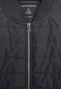 Černá voděodolná bunda s prošívaným vzorem, obsahující kovový zip a žebrovaný límec. Na štítku je uvedeno, že je vyrobena v Bangladéši.