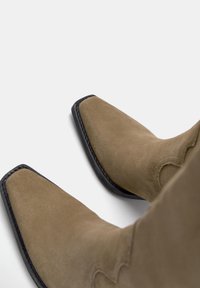 Brązowe zamszowe kowbojki z szpiczastym noskiem, stworzonymi z przeszyciami oraz czarną podeszwą. Gładka tekstura i elegancki design.