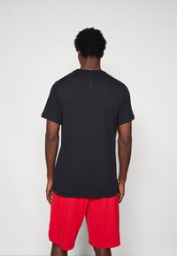 Μαύρο βαμβακερό T-shirt με κοντό μανίκι, στρογγυλή λαιμόκοψη και μικρή ετικέτα στην πλάτη. Συνδυασμένο με κόκκινες αθλητικές βερμούδες.