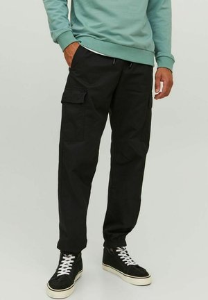 Mann trägt schwarze Cargohosen, schwarze High-Top-Sneaker mit weißen Schnürsenkeln und ein türkisfarbenes Sweatshirt, steht mit einer Hand in der Tasche.