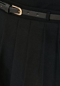 Musta laskostettu hame, jossa on teksturoitu kangas. Varustettu kapealla mustalla nahkavyöllä ja kultaisella soljella kiinnittämistä varten.