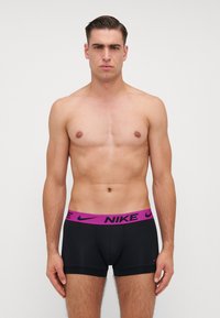 Czarne bokserki Nike z fioletowym pasem, na którym znajduje się logo. Model jest bez koszulki, przyjmuje neutralną pozę i wyraz twarzy.