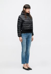 Γυναίκα που φοράει μαύρο κοντό μπουφάν-φουσκωτό, μπλε τζιν με ίσια ποδόγυρα και μαύρα φλατ παπούτσια, στέκεται μπροστά σε λευκό απλό φόντο.