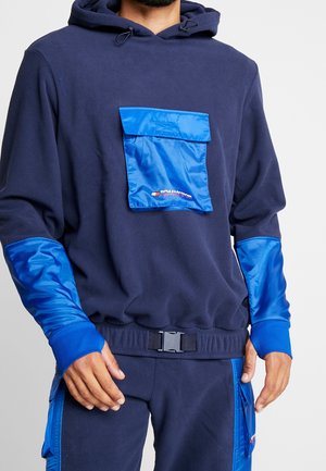 Man die een marineblauwe hoodie en broek draagt met felblauwe, glanzende zakken en elleboogstukken, een rits met gesp aan de voorkant en cargozakken.